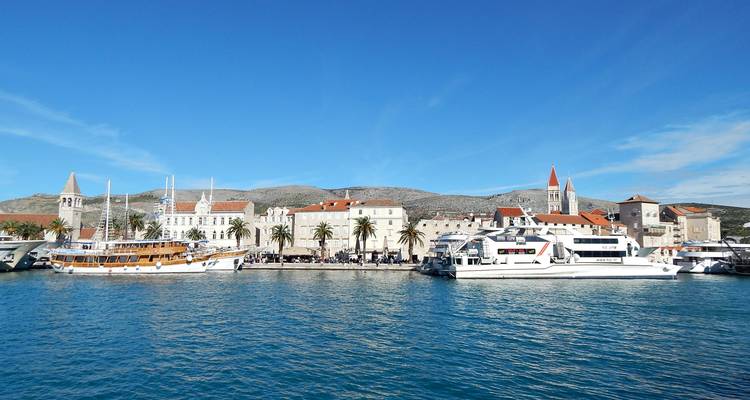 De waterkant van Trogir met palmbomen, historische torens en aangemeerde excursieboten in turquoise water.