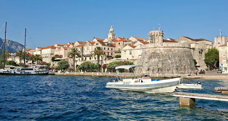 De stenen toren en middeleeuwse muren van de oude stad Korčula rijzen uit achter een kleine jachthaven op een zonnige dag.