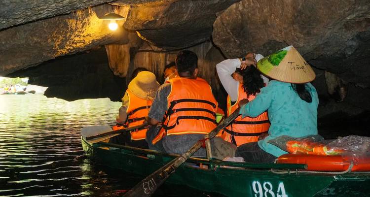 Tourists in a rowboat wearing orange vests enter a low limestone cave with dim lighting.