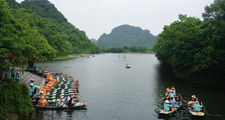 Rowboats with tourists line a dock on a scenic river surrounded by lush karst hills.