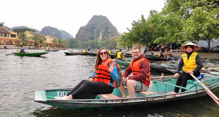 Tourists in metal sampan smile and wave on calm water with karst backdrop.