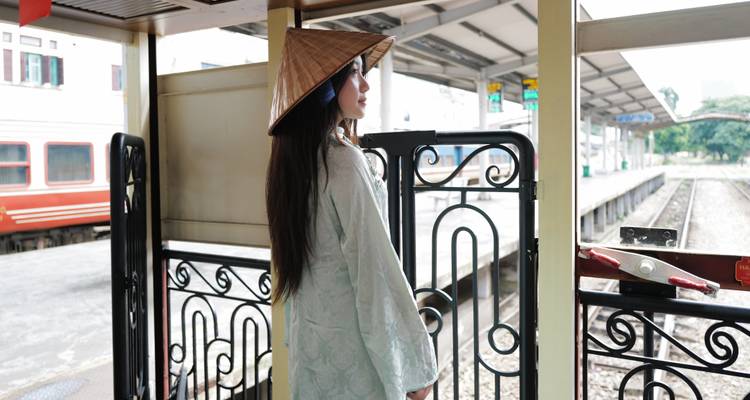 Eine Frau, die einen traditionellen kegelfoermigen Hut traegt, steht auf einem Bahnsteig im Vintage-Stil und blickt auf die Gleise hinunter.