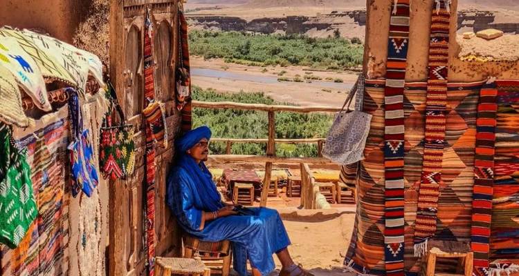 Een man in een blauwe djaellaba die ontspant tussen kleurrijke Berber tapijten met een woestijnvallei op de achtergrond in Ait Benhaddou.