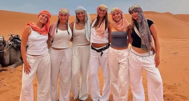 Zes vrouwen in gecoördineerde woestijnoutfits die op oranje duinen staan met een kameel