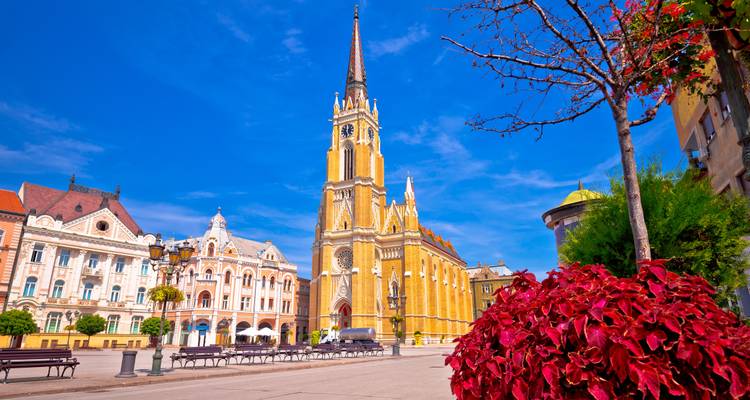 Liberty Square in Novi Sad with the ornate yellow Name of Mary Church tower under a vivid blue sky and red flowers in foreground.
Freiheitsplatz in Novi Sad mit dem verzierten gelben Turm der Marienkirche unter einem leuchtend blauen Himmel und roten Blumen im Vordergrund.