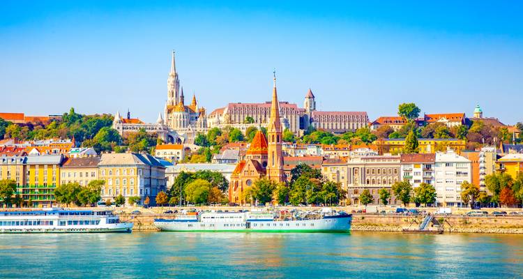 Des bâtiments historiques colorés et des clochers d'églises s'élèvent au-dessus du Danube par une belle journée ensoleillée, avec des bateaux amarrés le long de l'embarcadère.