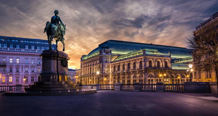 Vue élégante en soirée de l'Opéra d'État de Vienne avec une statue équestre en avant-plan sous un ciel au coucher de soleil dramatique.