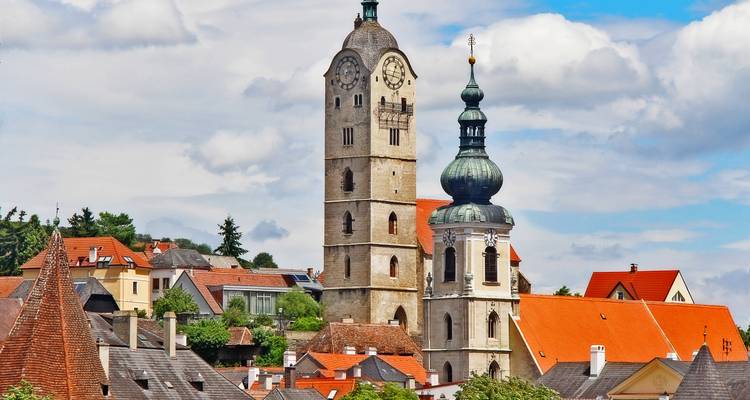 Panorama ensoleillé d'une ville autrichienne historique avec deux clochers d'églises distinctifs s'élevant au-dessus des toits en tuiles rouges.