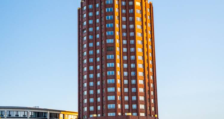 Close-up van de bakstenen en glazen gevel van de Westend Tower in Frankfurt, badend in warm ochtendlicht tegen een strakblauwe hemel.