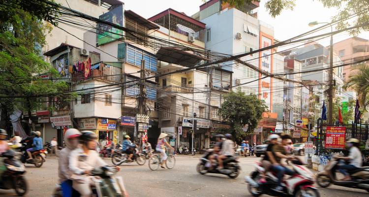 Chaotic Hanoi street scene with blurred motorbikes criss-crossing beneath tangles of overhead power lines.