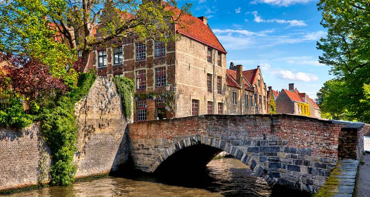 Un pont de pierre s'arque au-dessus d'un canal bordé d'arbres à côté de maisons médiévales en brique à Bruges sous un ciel bleu.