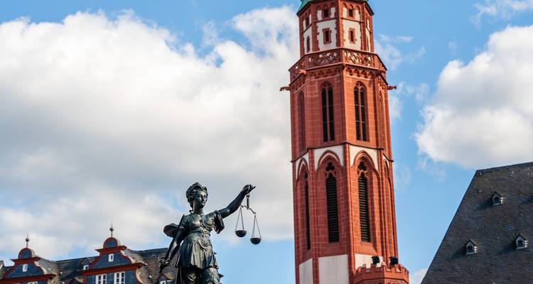 Statue de bronze de la Dame Justice encadrée par une tour d'église rouge sous un ciel bleu à Francfort.