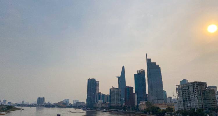 Modern Ho Chi Minh City skyline lining the Saigon River with the sun glowing through hazy sky.