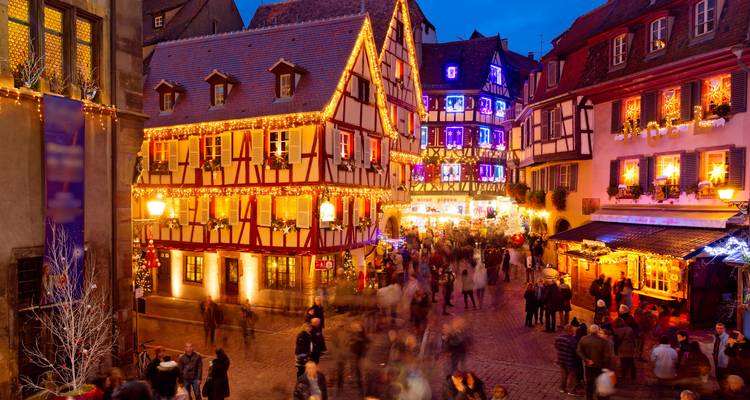 Abendliche Szenerie von Fachwerk-Gebäuden in Colmar, die in Weihnachtslicht erstrahlen und von geschäftigen Menschenmengen belebt werden
