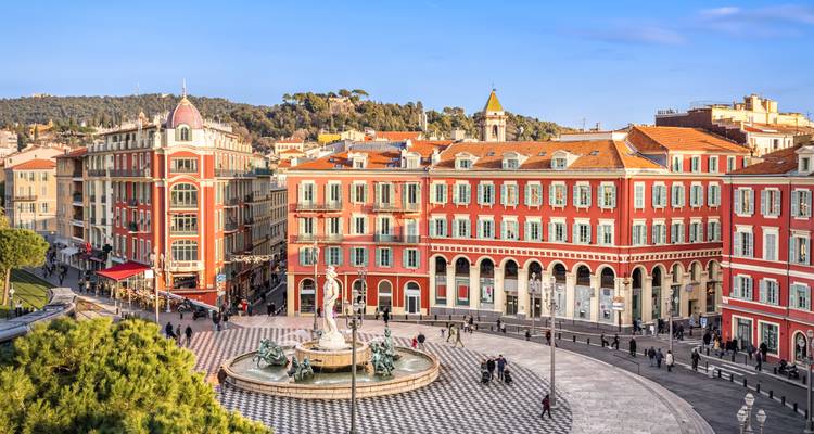 La Place Masséna à Nice avec ses arcades aux façades rouges, sa fontaine centrale, et les piétons profitant d'un après-midi ensoleillé.