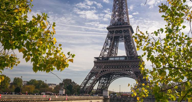 Vue latérale de la Tour Eiffel encadrée par des feuilles d'automne sous des nuages striés lors d'un après-midi dégagé.