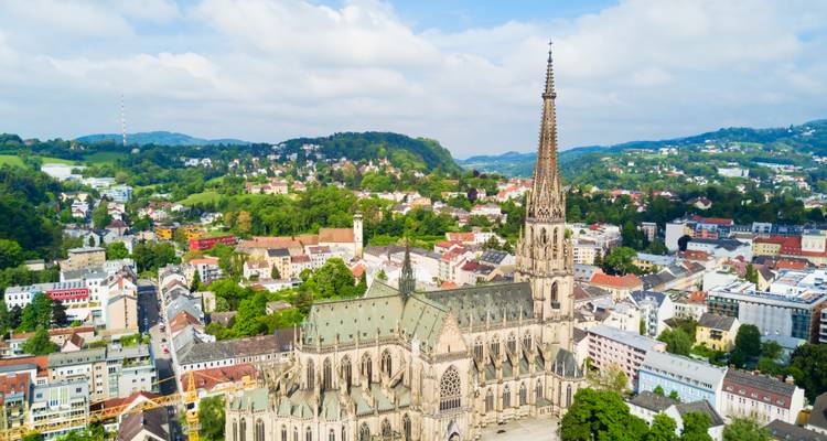 Aerial view over Linz featuring the towering spire of the New Cathedral amid a patchwork of colorful city blocks and green hills.
