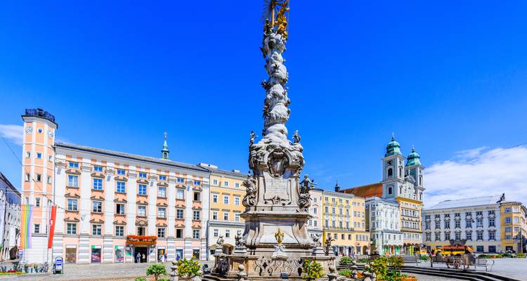 Vibrant main square of Linz with the ornate Trinity Column against a deep blue sky and pastel-colored facades lining the plaza.