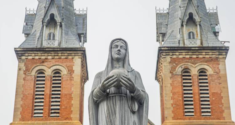 Estatua de la Virgen María en primer plano con las dos agujas de la Basílica Catedral de Notre-Dame de Saigón de fondo contra un cielo nublado.