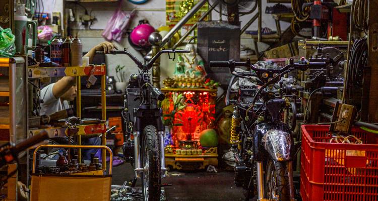 Taller de reparación de motos desordenado en Vietnam con el brazo de un mecánico extendido y un pequeño santuario rojo de fondo.