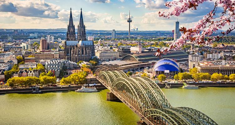 Panorama de Colonia con el Puente Hohenzollern y la catedral de dos torres enmarcados por flores de cerezo.