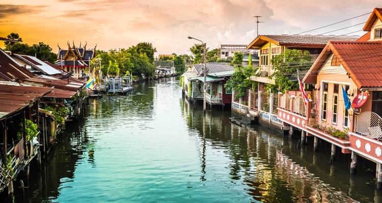 Peaceful canal lined with traditional wooden houses and small temples in Bangkok at golden hour.