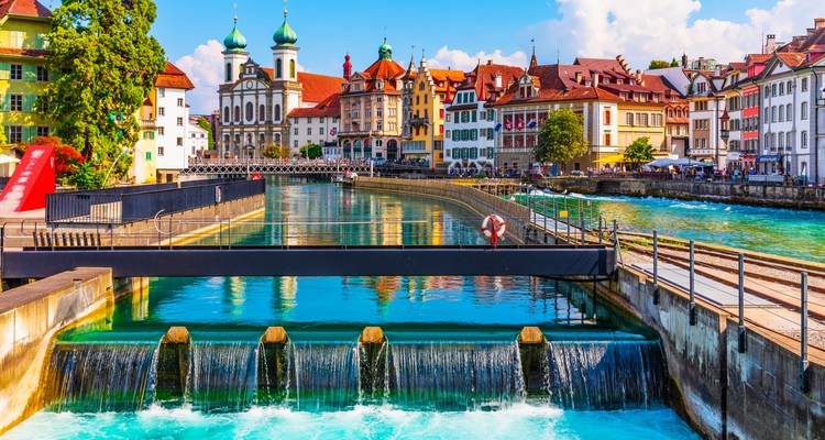 Schilderachtige rivierpromenade met barokke kerktorens en turquoise water dat over een stuw stroomt.