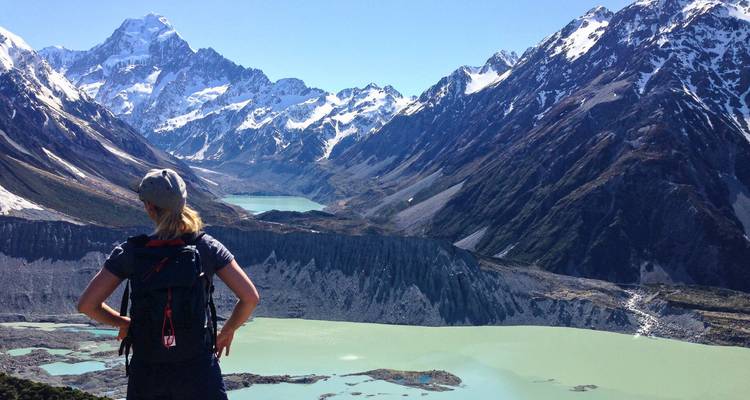 Wandelaar staat met rugzak en kijkt uit over gletsjermeren en sneeuwbedekte bergen van Aoraki/Mount Cook National Park.