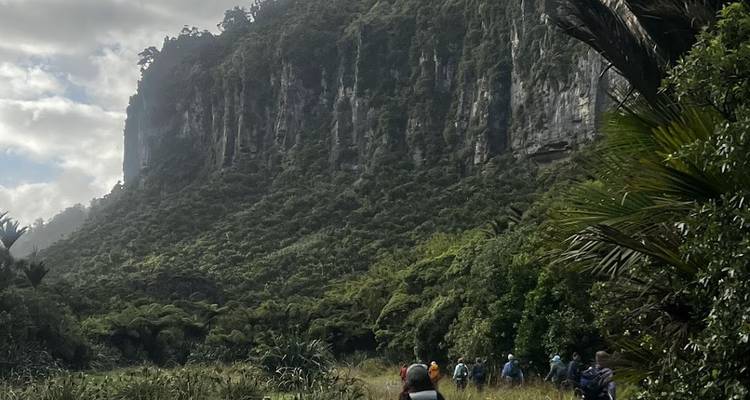 Wandelgroep loopt door een weelderig groene vallei onder een imposante rotsformatie die bedekt is met jungle-begroeiing.