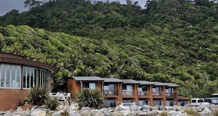Houten lodge complex verscholen tussen dicht kustbos met geparkeerde busjes op rotsachtig strand.