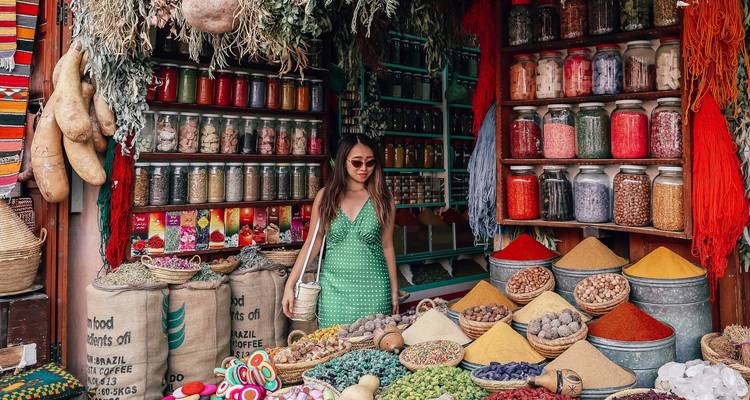 Puesto de especias marroquí lleno de color con conos vibrantes, frascos y hierbas colgantes, un comprador mirando.