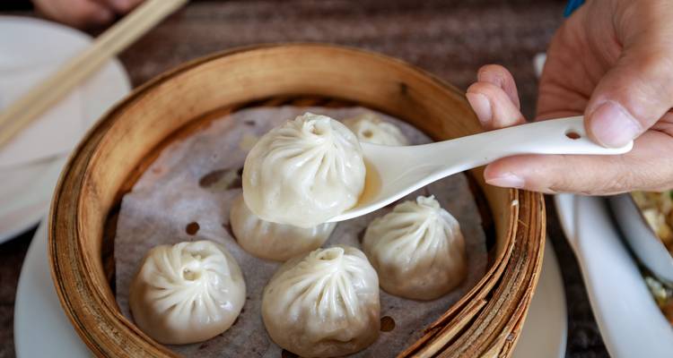Close-up van dampende xiao long bao dumplings, waarbij er een uit het bamboe mandje wordt opgetild met een keramische lepel.