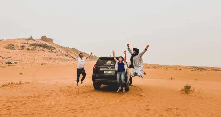 Drie reizigers springen blij naast een 4x4-voertuig te midden van enorme oranje woestijnduinen.