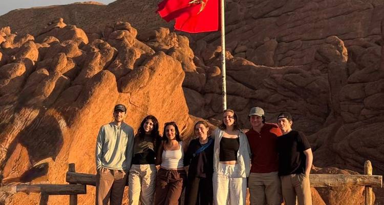 Groep reizigers poseert onder de Marokkaanse vlag op rode rotsformaties in het licht van de late middag.