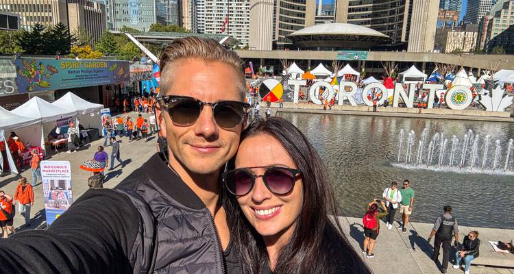 Een glimlachend stel maakt een selfie met uitzicht op Torontos Nathan Phillips Square tijdens een levendig buitenevenement.