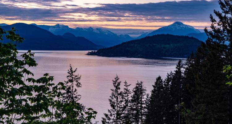 Vue au crépuscule sur un littoral bordé d'arbres et des îles montagneuses en silhouette sous un ciel aux couleurs variées.