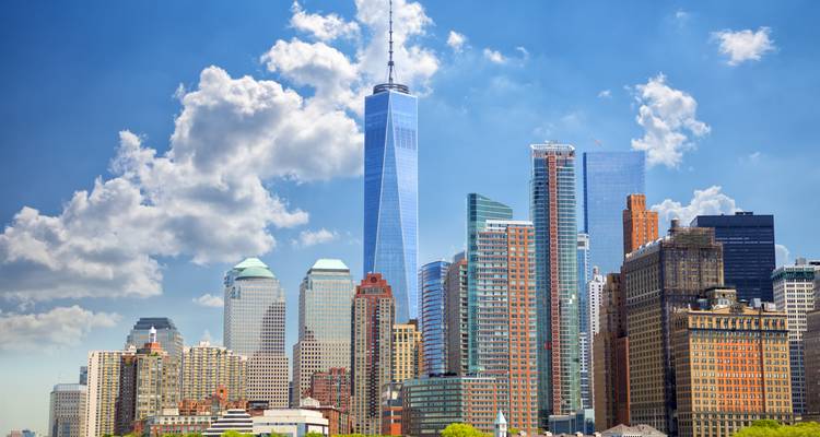 Bright midday shot of Lower Manhattan skyline dominated by One World Trade Center and cumulus clouds above.