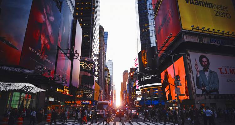 Bustling Times Square intersection at sunset with glowing billboards and crowds crossing.