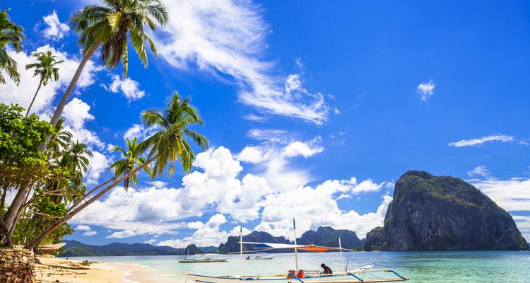 Plage tropicale bordée de palmiers avec un bateau à balancier et une impressionnante île calcaire sous un ciel d'un bleu éclatant.