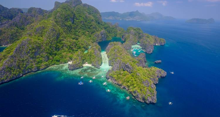 Vue aérienne d'îles karstiques verdoyantes et accidentées entourées d'une mer bleu profond parsemée de petits bateaux.