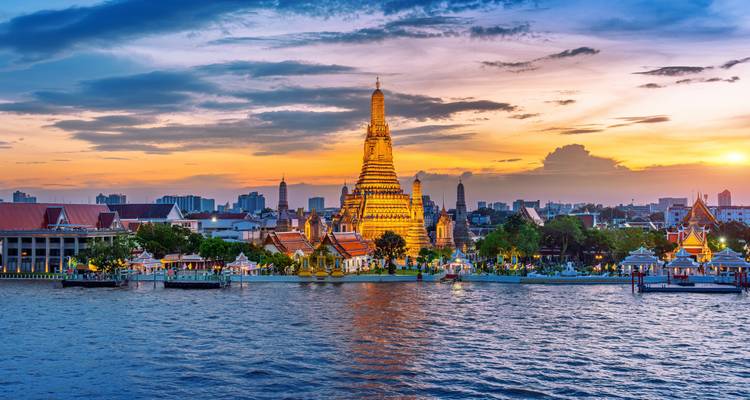 Le magnifique temple Wat Arun brille d'une teinte dorée au coucher du soleil de l'autre côté du fleuve Chao Phraya avec la skyline de la ville en arrière-plan.