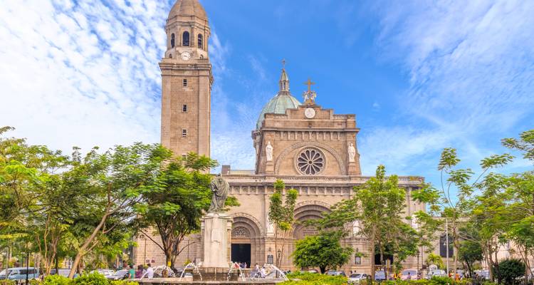 La Cathédrale de Manille se dresse derrière des arbres luxuriants et une statue historique sous un ciel bleu éclatant.