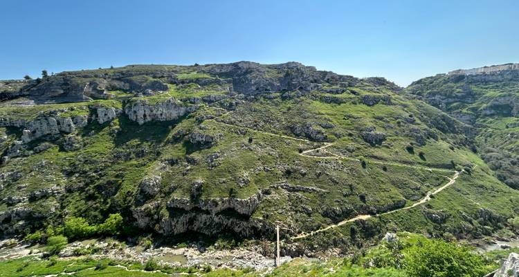 Uitgestrekte groene ravijn met kronkelig wandelpad en kalksteenrotsen onder een heldere blauwe lucht in de buurt van Matera