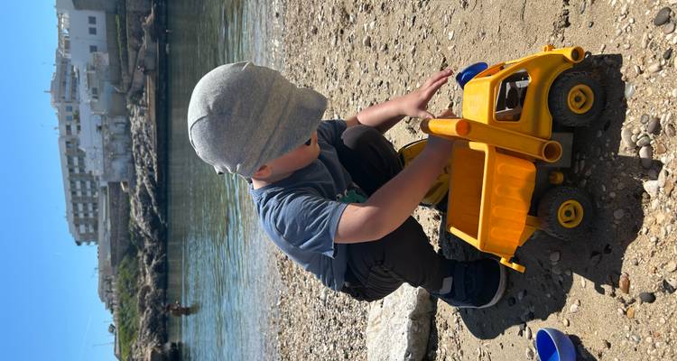 Jongetje dat speelt met een speelgoedstortwagen op een klein kiezelstrand naast een rustige baai