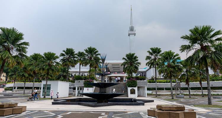 Place de mosquée moderne avec fontaine géométrique, palmiers et minaret élancé sous un ciel gris à Kuala Lumpur