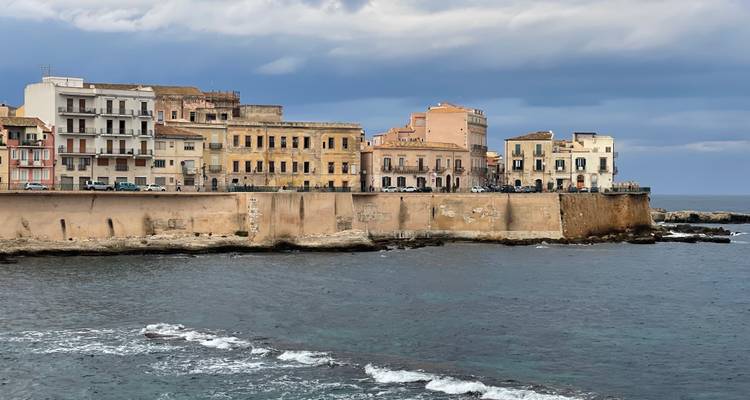 Vue du front de mer des anciens bâtiments fortifiés d'Ortigia s'étendant dans la mer Ionienne sous un ciel nuageux.
