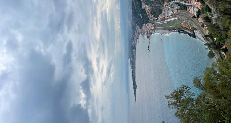 Vue panoramique élevée sur un littoral sinueux et une ville côtière avec une ligne ferroviaire et des eaux turquoise sous des nuages spectaculaires.