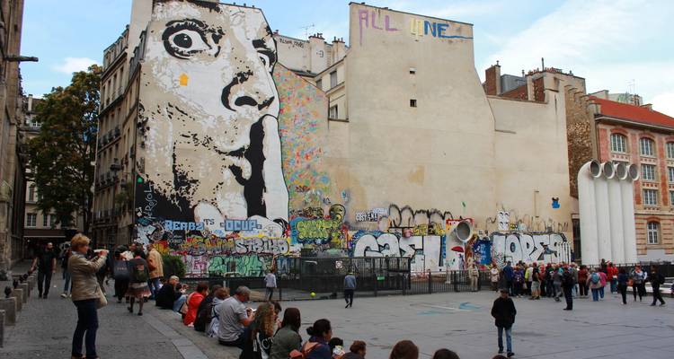 Grande fresque murale de graffiti représentant un visage faisant signe de se taire sur un mur de bâtiment près du Centre Pompidou avec des foules assises en dessous.