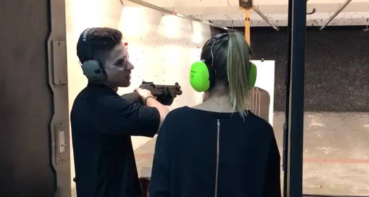 Un instructeur guide une femme dans un stand de tir couvert; tous deux portent une protection auditive tout en visant un pistolet.