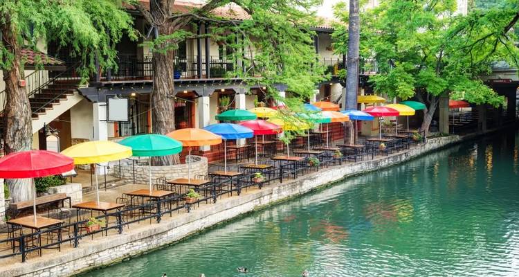 Tables de café fluviales dynamiques aux parasols multicolores le long de la promenade turquoise de la rivière San Antonio.